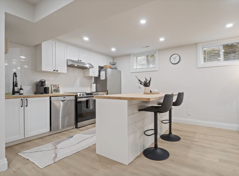 Lower-level kitchen with white shaker cabinets, butcher-block peninsula with two stools and stainless-steel appliances