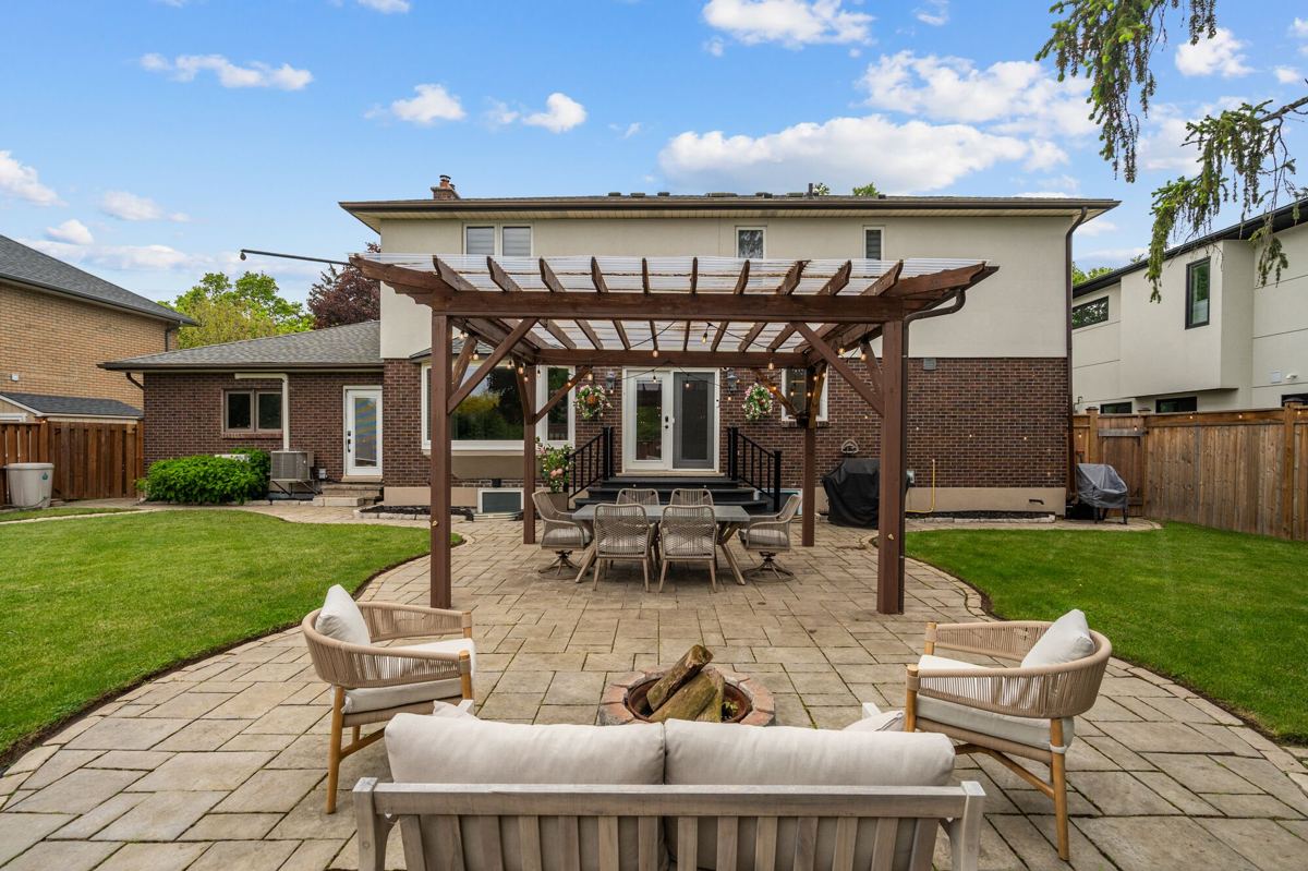 Rear of 7 Carl Crescent: a two-storey brick and stucco home with a wood pergola over a stone patio, a dining set, fire pit seating and lawn